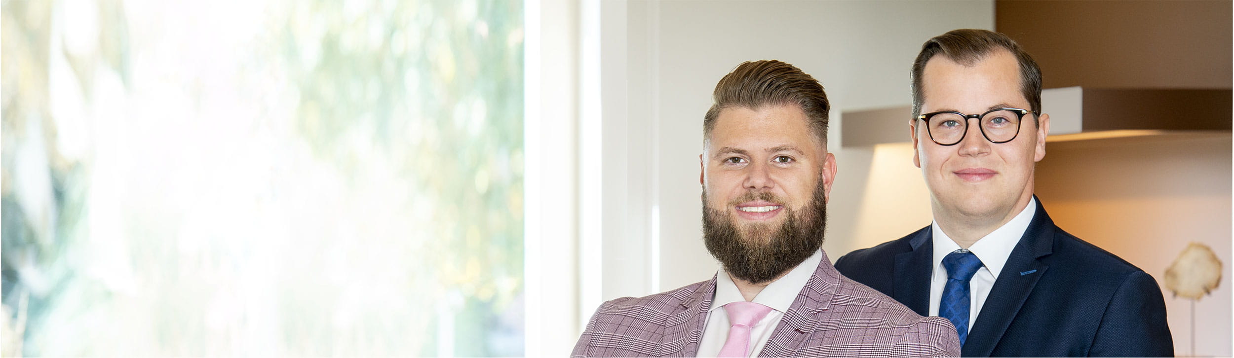 Twee personen in formele kleding staan naast elkaar binnen in een lichte ruimte, met een raam en onscherpe groene achtergrond.